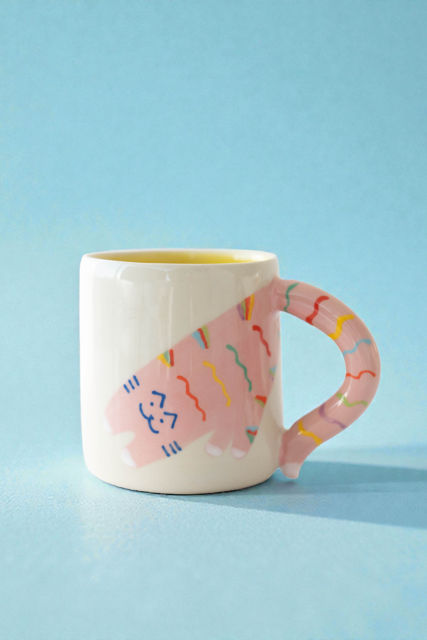 White mug with pink handle and colorful cat design on a light blue background
